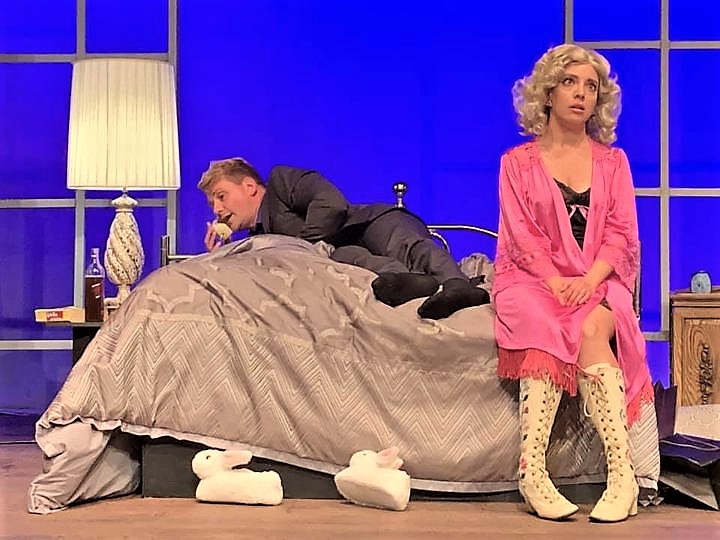 Photo of set on stage. Man in suit lays out on the bed holding a phone to his ear. Woman sits on edge of bed in robe and possibly lingerie underneath with knee high laced boots. 